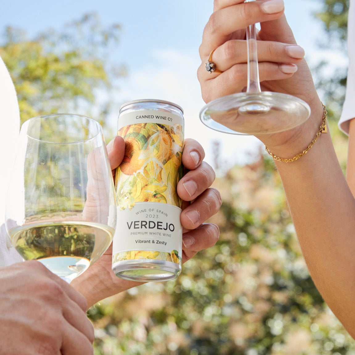 Two people holding glasses of white wine and a bottle labeled 'Verdejo' against a blurred natural background.